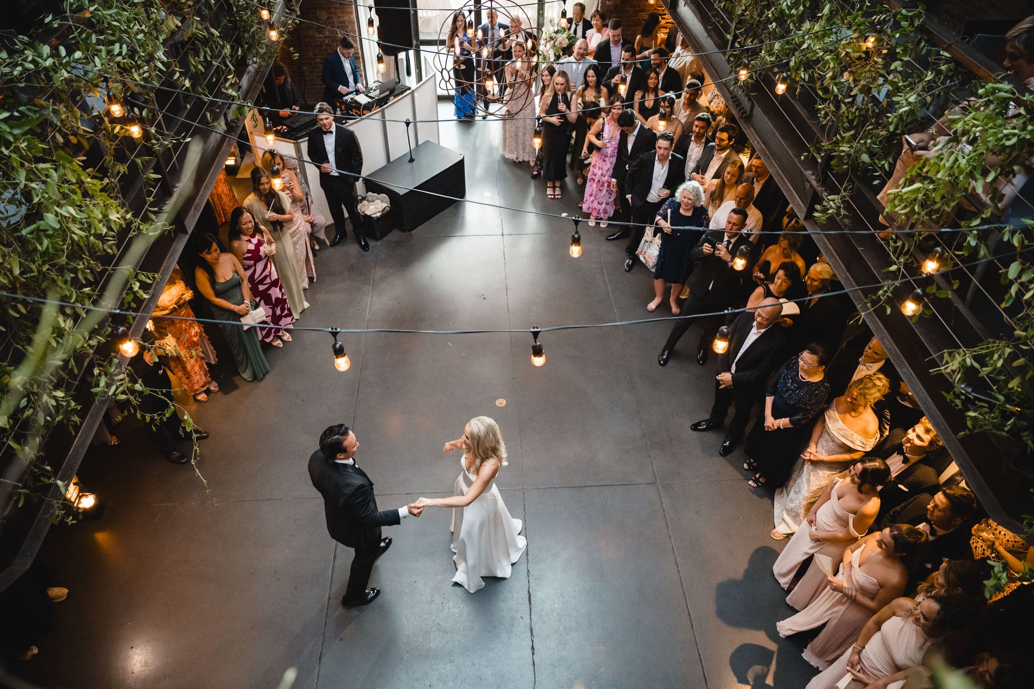 Aerial view of bride and groom sharing first dance surrounded by guests at an indoor wedding venue.