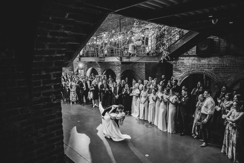 Black and white photo of bride and groom sharing first dance at wedding reception with guests watching.