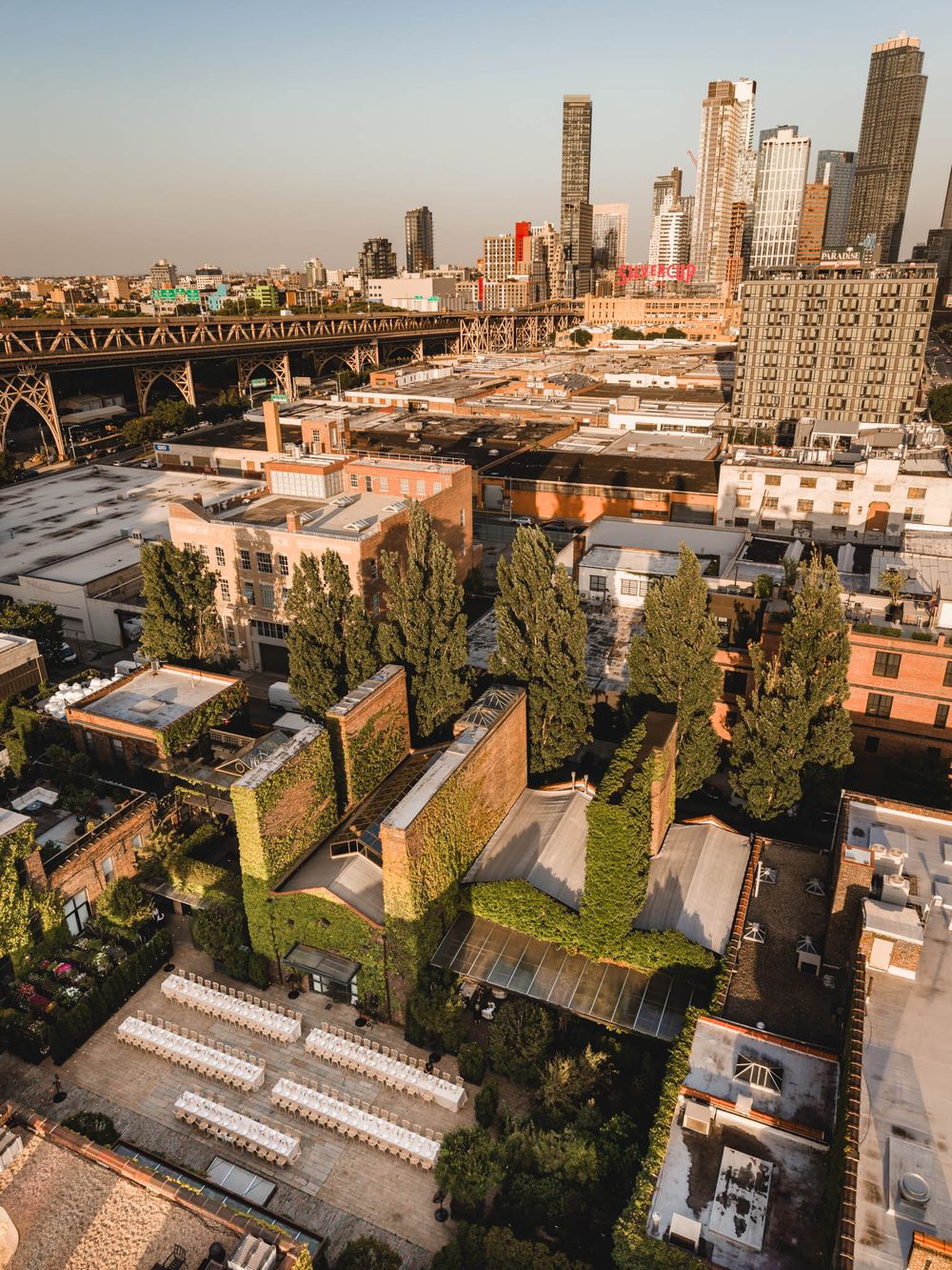 Aerial view of green-covered brick buildings with rooftop gardens near a bridge, with NYC skyline at golden hour.