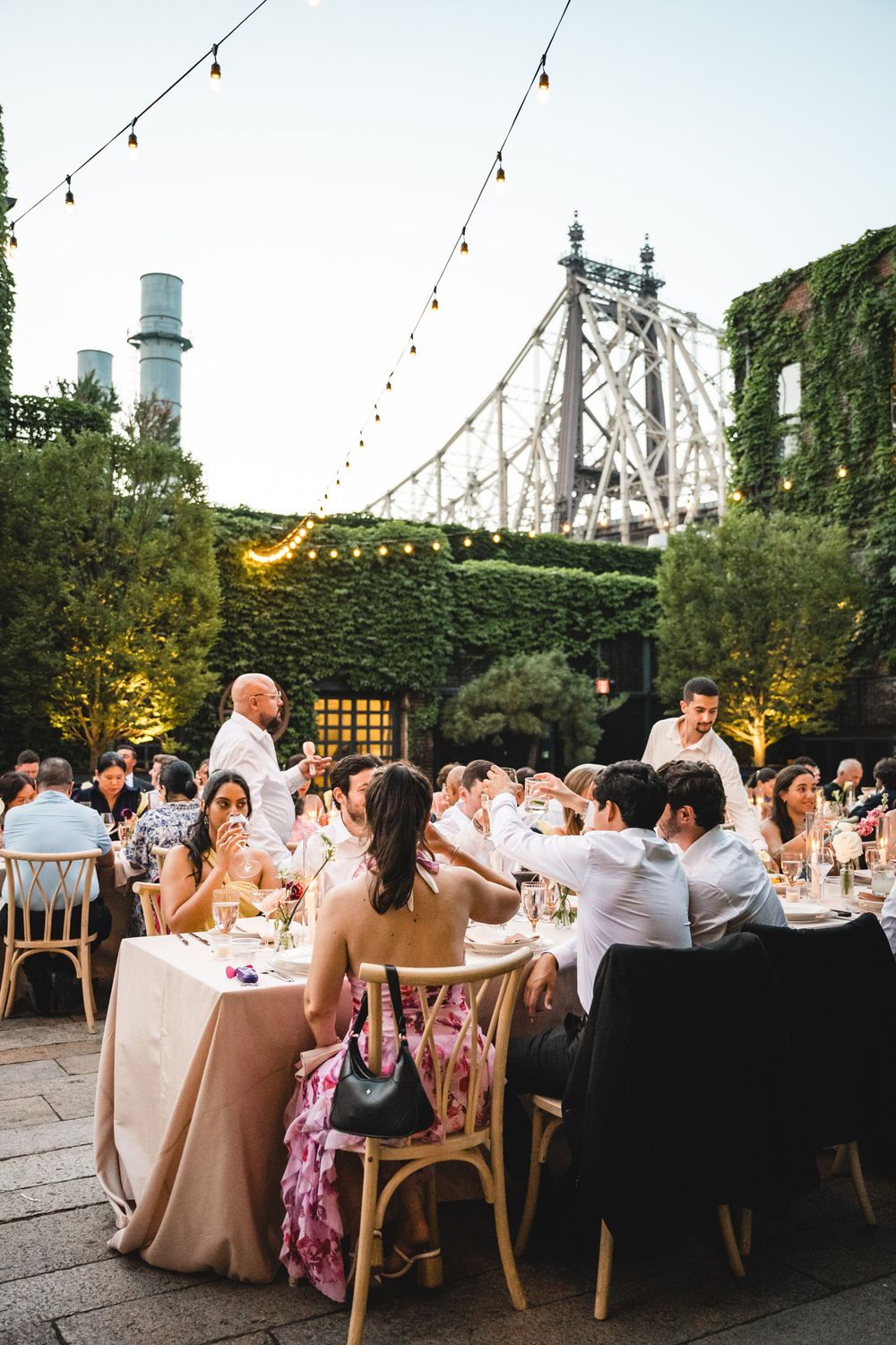 Outdoor wedding reception dinner under string lights with Queensboro Bridge backdrop in New York City.