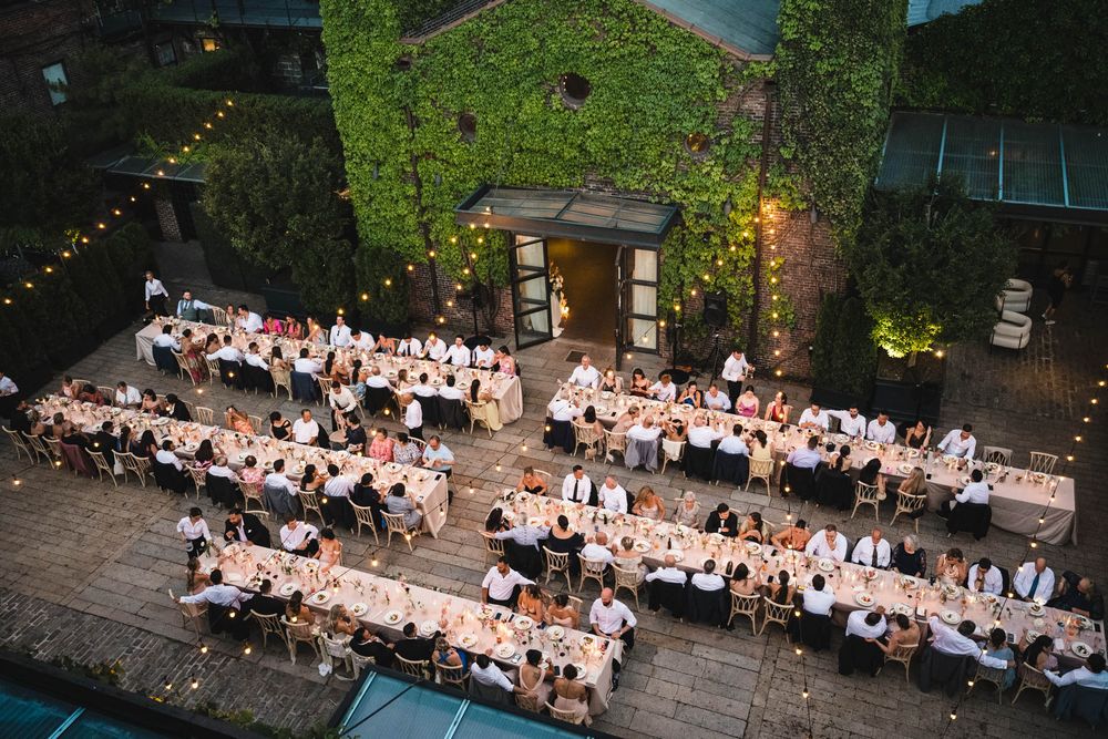 Aerial view of an elegant outdoor wedding reception with long tables, string lights, and ivy-covered venue at dusk.