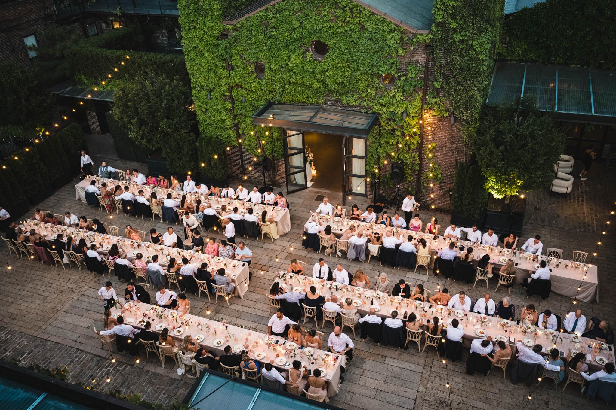 Aerial view of an elegant outdoor wedding reception with long tables, string lights, and ivy-covered venue at dusk.