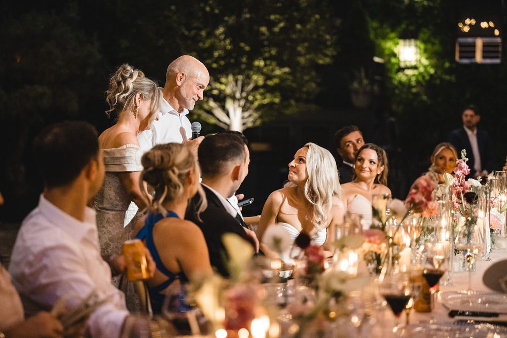 Guests laugh and celebrate at an elegant outdoor wedding reception dinner with candlelit tables and floral arrangements.