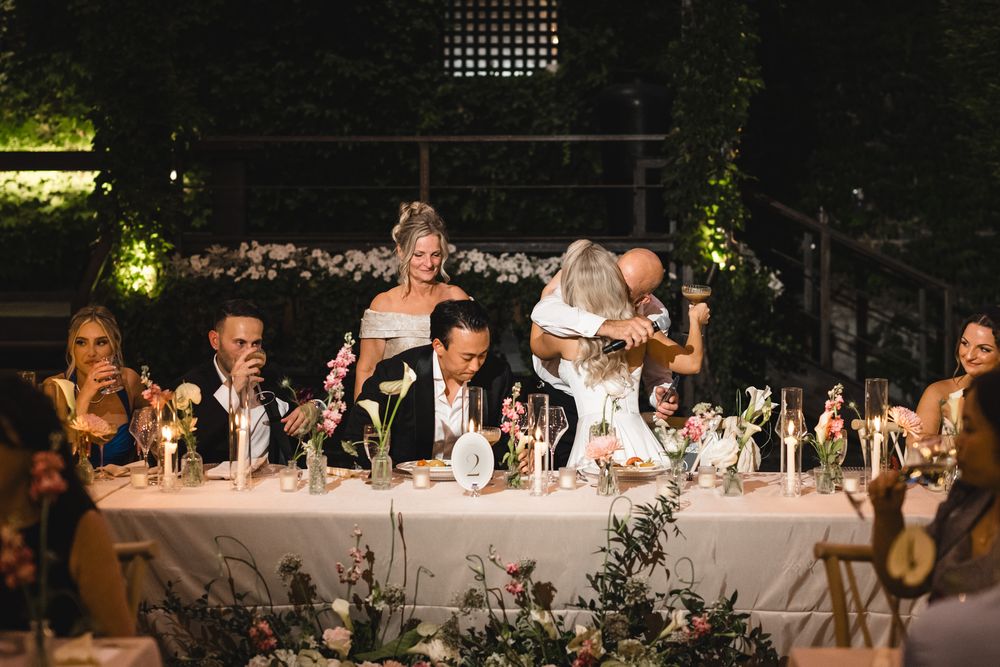 Wedding party at head table during reception with couple sharing a moment under soft evening lighting.