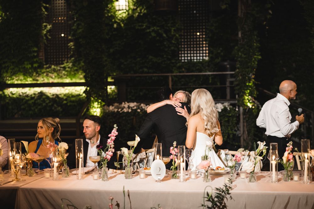Bride and groom share a heartfelt embrace at their decorated wedding reception head table during speeches.