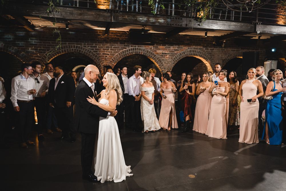 Bride and groom share first dance in rustic brick arch venue surrounded by wedding party guests.