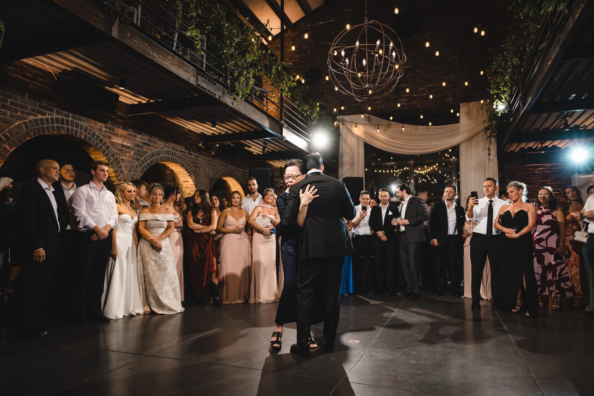 Elegant wedding reception in a brick venue with string lights, guests watching couple dance on the floor.