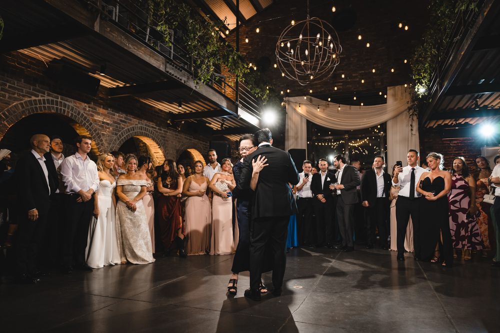 Elegant wedding reception in a brick venue with string lights, guests watching couple dance on the floor.