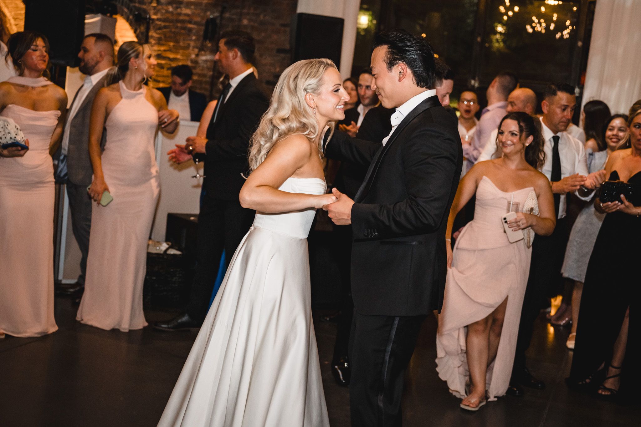 Bride in white gown and groom in black tuxedo share their first dance surrounded by wedding guests.