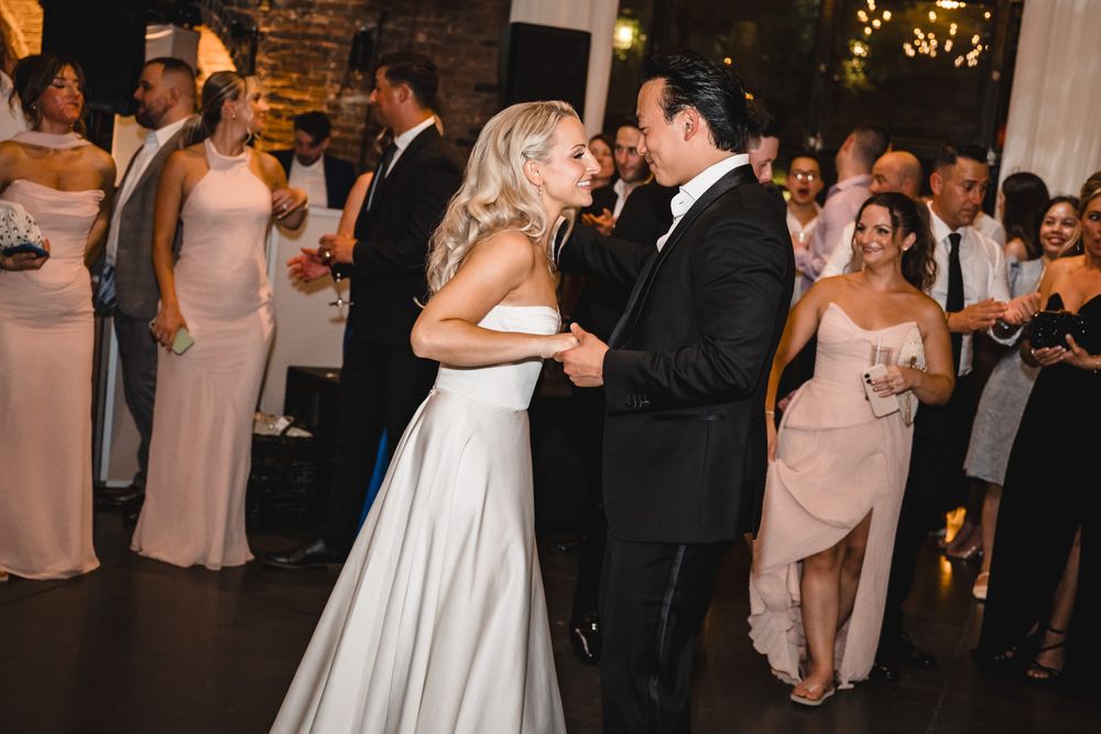 Bride in white gown and groom in black tuxedo share their first dance surrounded by wedding guests.