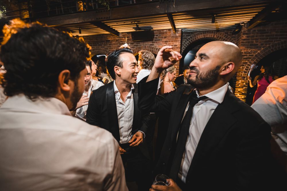 Men in suits socializing and laughing at an indoor party event with brick walls and dim lighting.