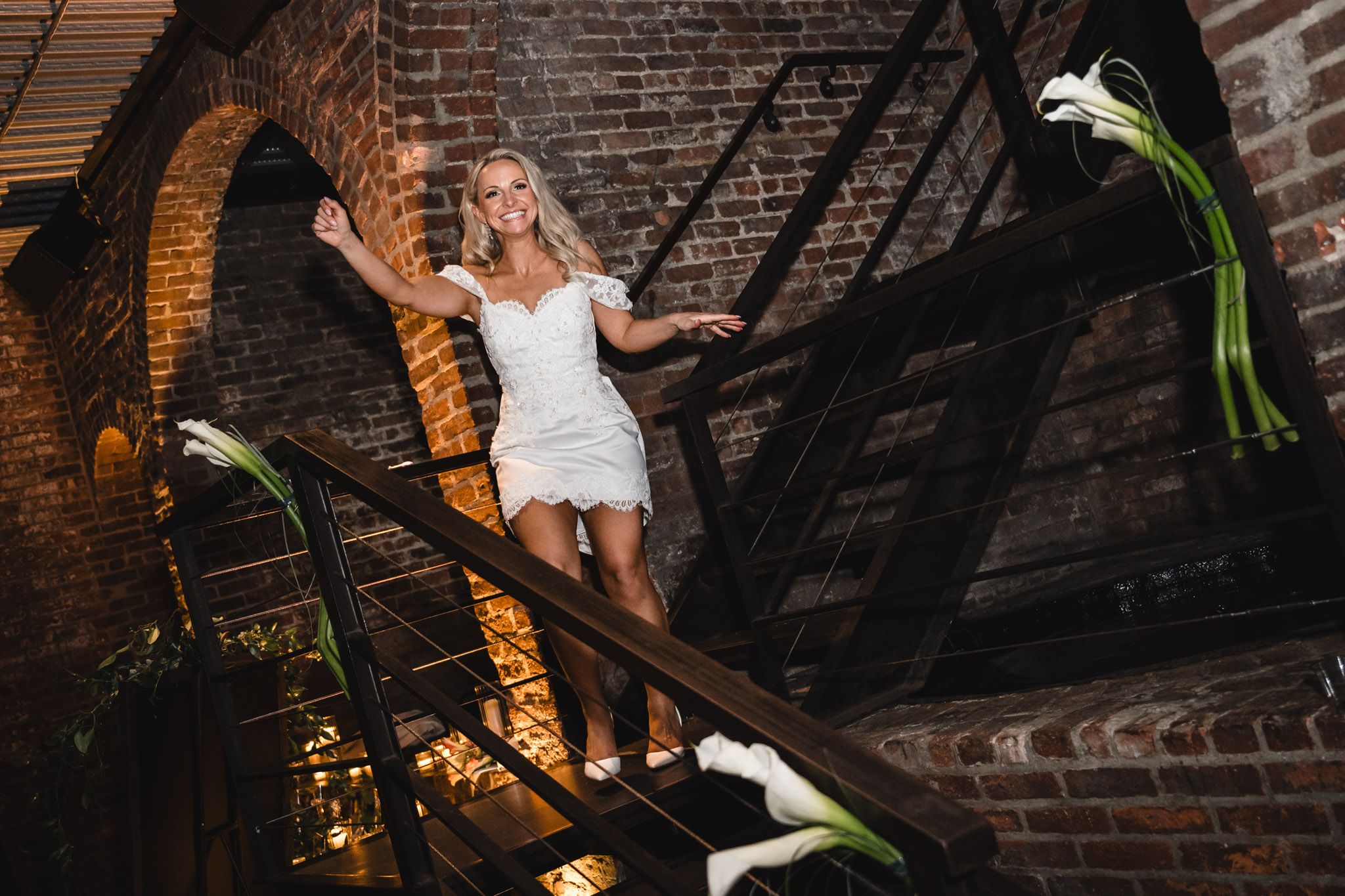 Blonde bride in short white dress posing on rustic brick staircase with white calla lilies.