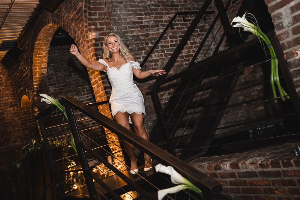 Blonde bride in short white dress posing on rustic brick staircase with white calla lilies.