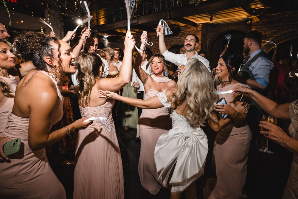 Energetic wedding reception dance floor with bride, groom, and bridesmaids in blush dresses celebrating.