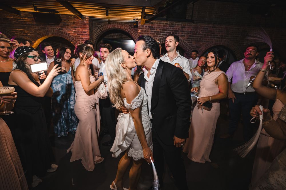 Bride and groom share a passionate kiss on the dance floor surrounded by cheering wedding guests.