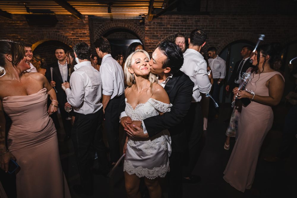 Bride and groom share a romantic kiss on the dance floor surrounded by wedding guests during reception.