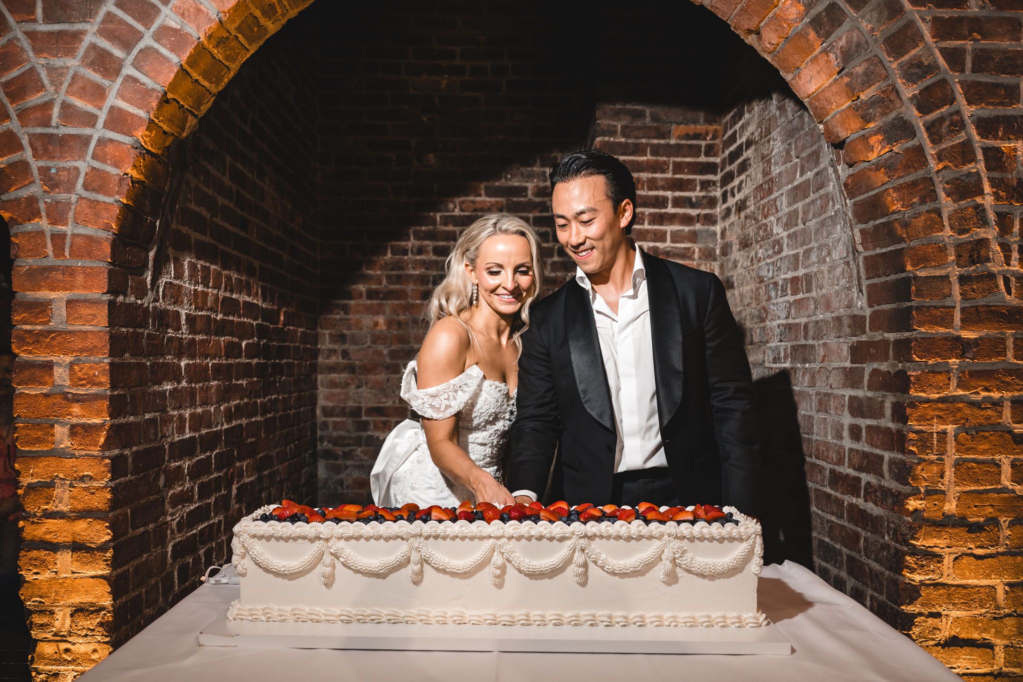 Bride and groom cut a large rectangular wedding cake decorated with strawberries in a brick archway.