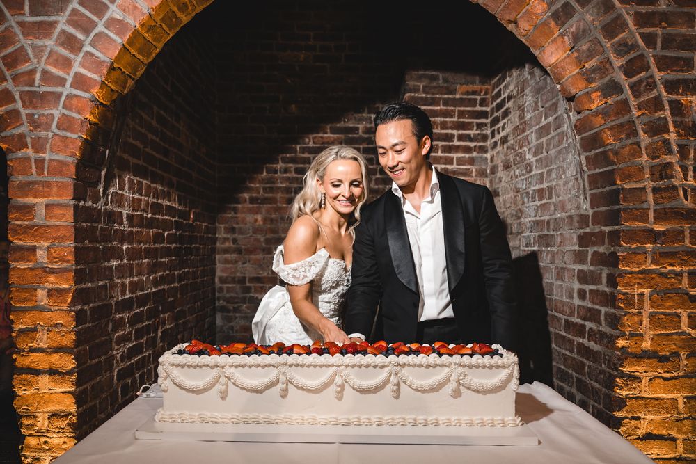 Bride and groom cut a large rectangular wedding cake decorated with strawberries in a brick archway.