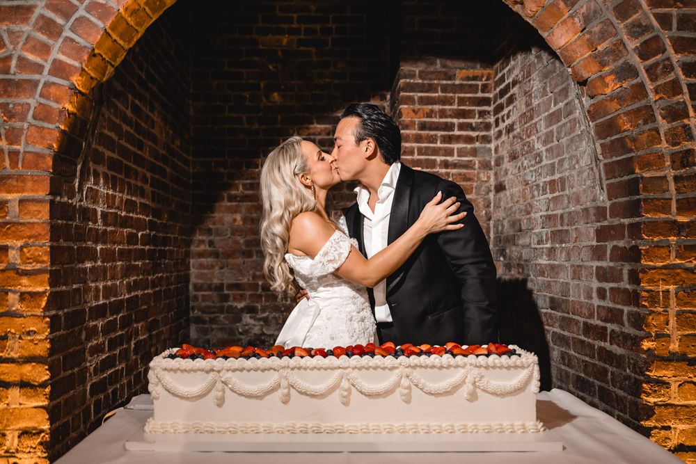 Bride and groom sharing a kiss behind a large white wedding cake in a brick archway.