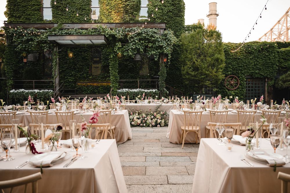 Elegant outdoor wedding reception with ivory-draped tables, floral centerpieces, and ivy-covered walls under string lights.
