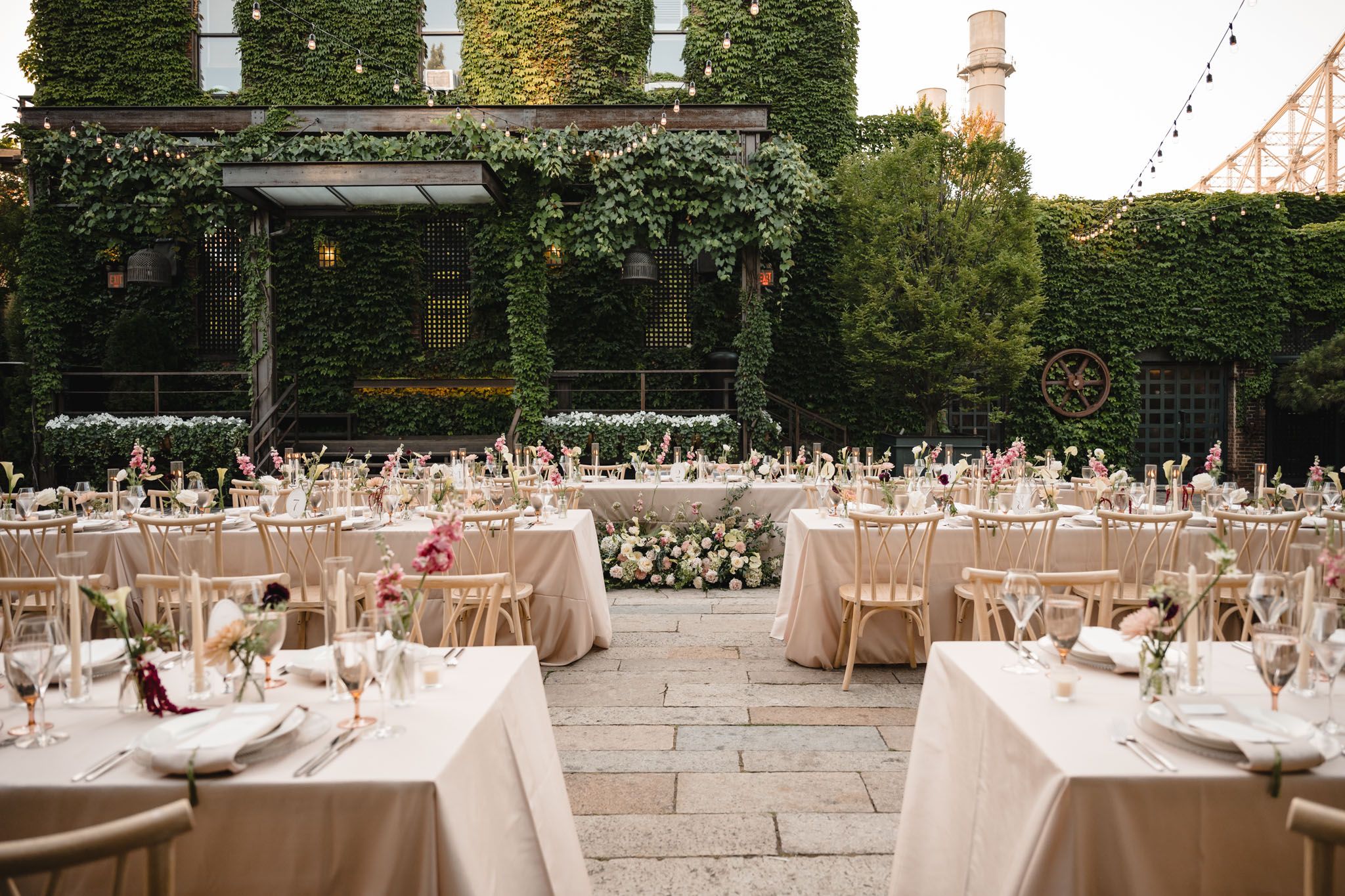 Elegant outdoor wedding reception with ivory-draped tables, floral centerpieces, and ivy-covered walls under string lights.