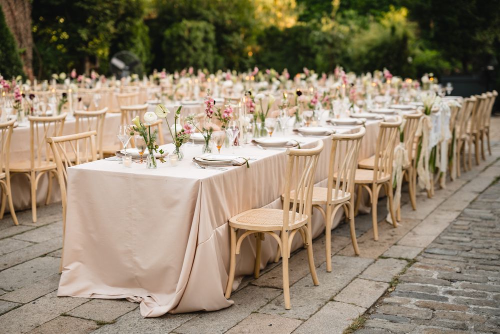 Elegant outdoor wedding reception with long tables draped in blush linens, wooden crossback chairs, and floral centerpieces.