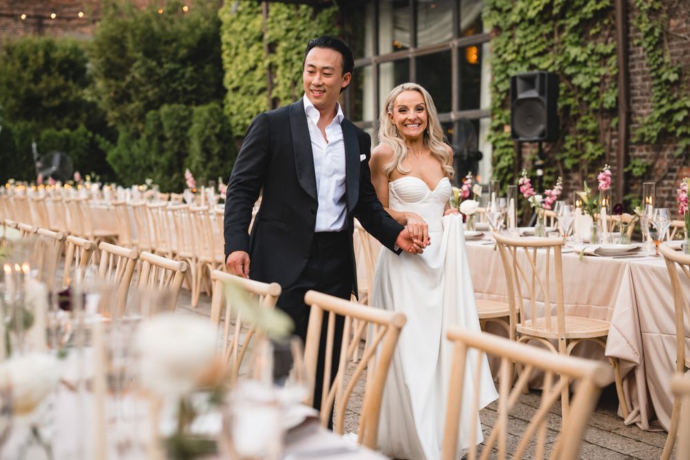 Bride and groom walk hand in hand through beautifully decorated outdoor wedding reception with long dining tables.