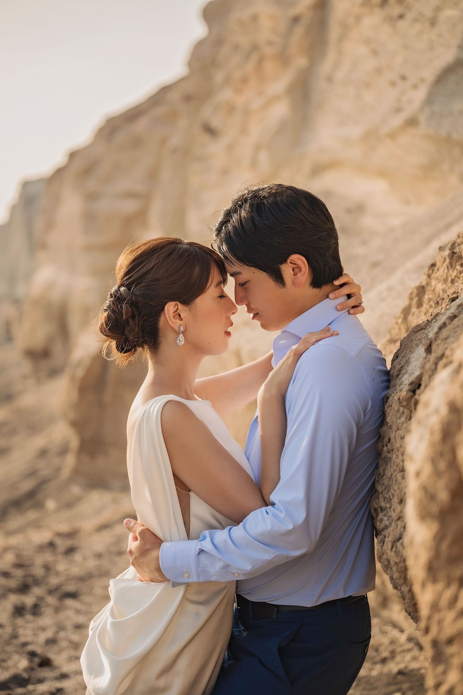 Couple in romantic embrace near sandy cliffs, woman in white dress with updo, man in blue shirt, pre-wedding photo shoot.