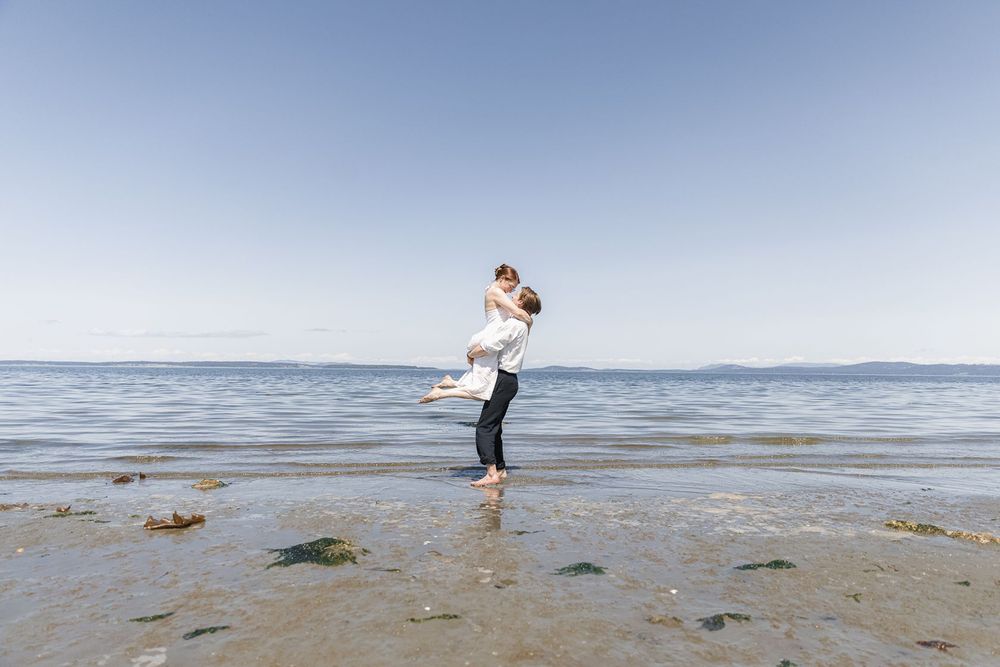 Couple embracing on a serene beach, man lifting woman joyfully with calm ocean waters and clear blue sky behind them.