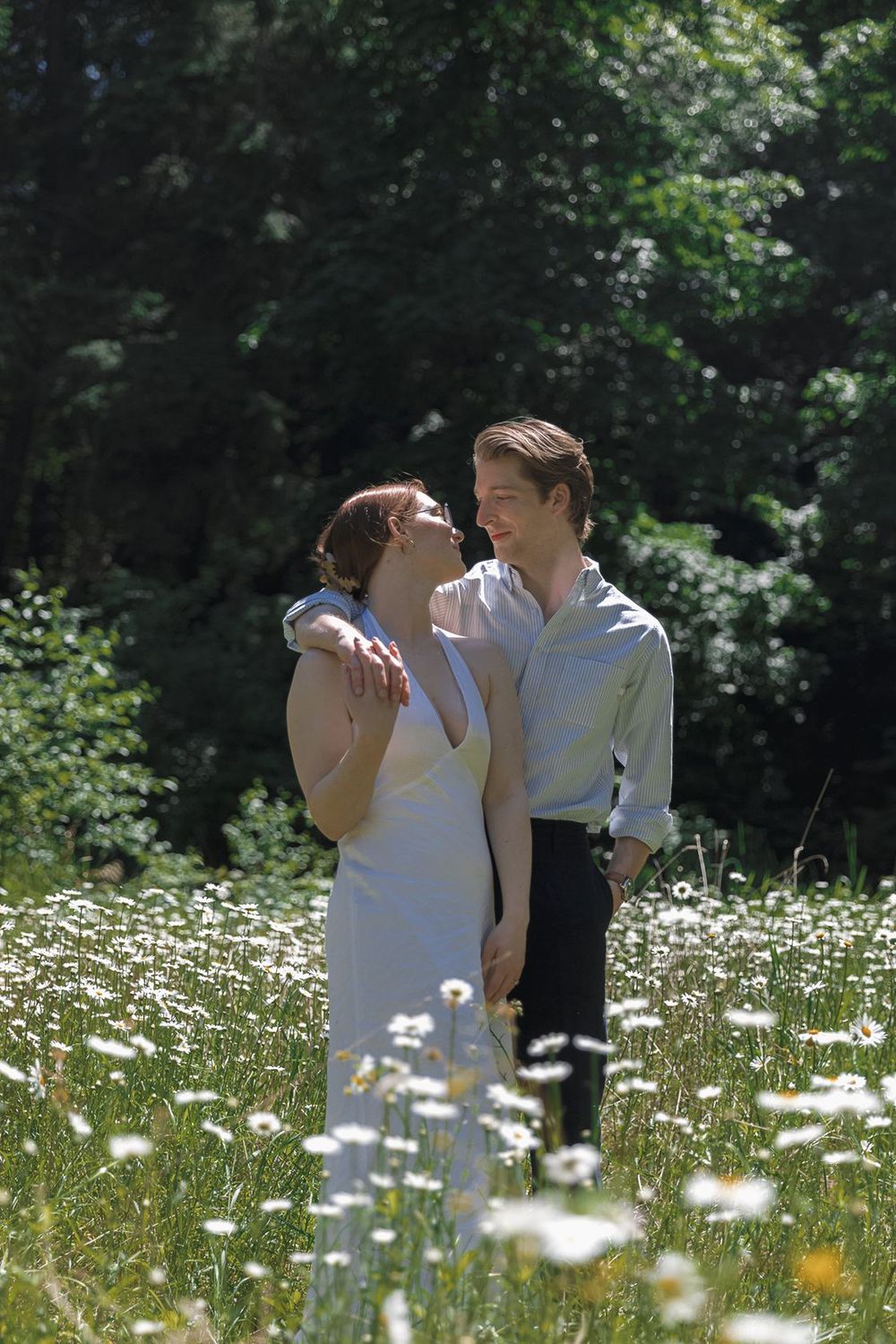 Couple embracing in a sunny wildflower meadow with white daisies, surrounded by lush green trees.