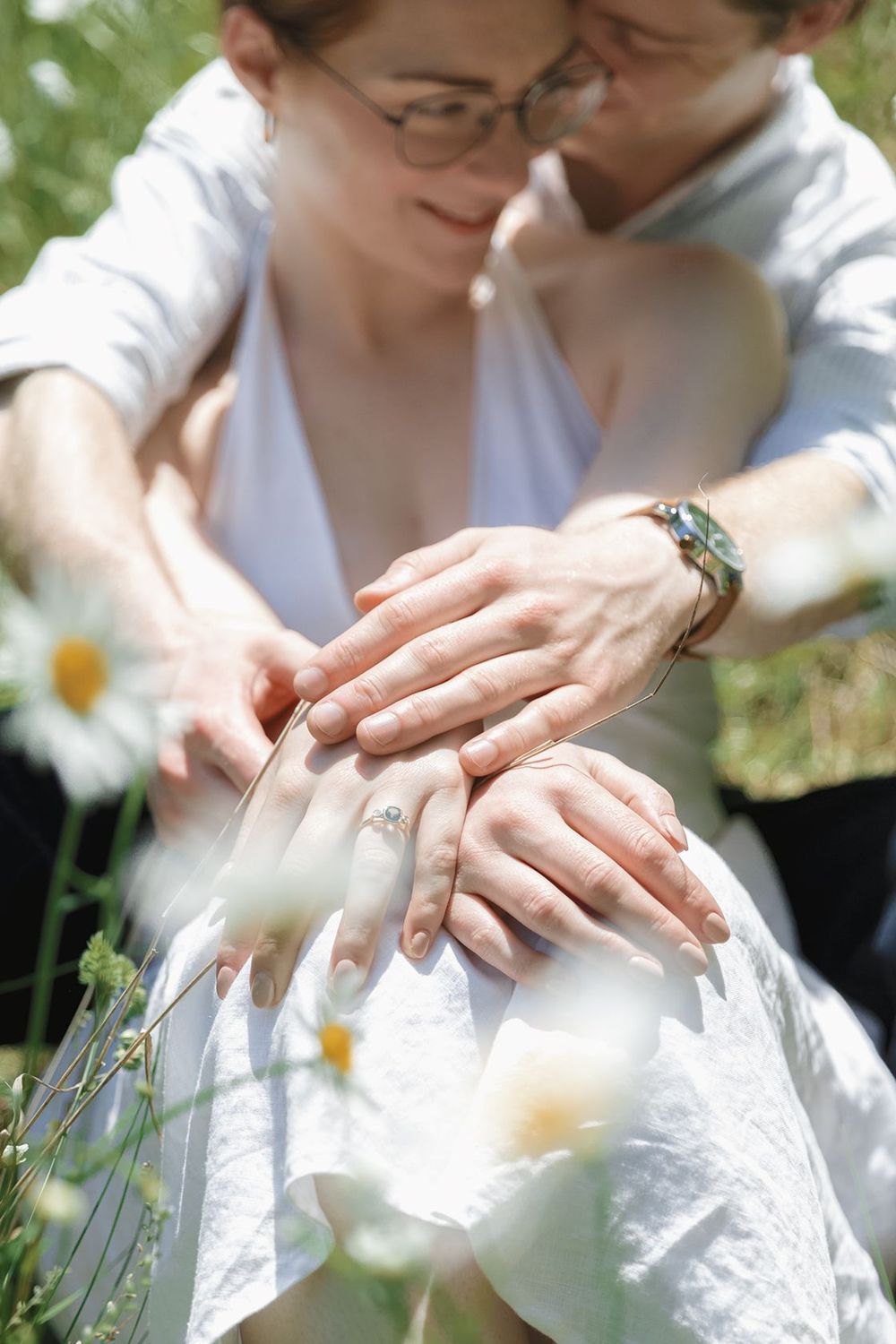 Couple sitting in a daisy field, hands intertwined showing wedding rings, wearing white and casual attire.