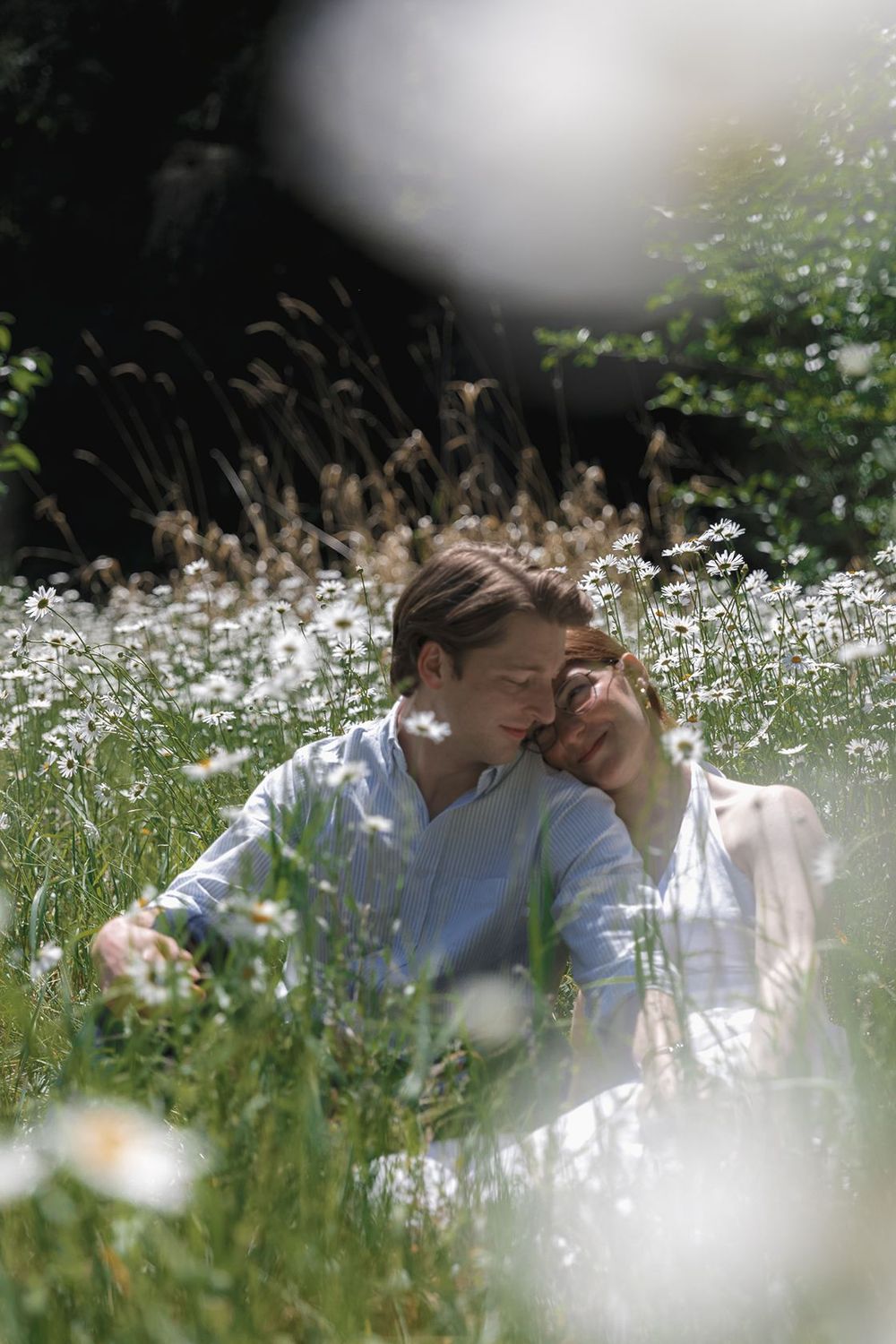 Couple sitting together in a sunny wildflower meadow with white daisies, sharing a tender romantic moment outdoors.