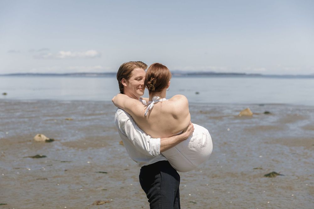 A groom lifts his bride in a romantic beach embrace, with calm ocean waters and a clear sky behind them.