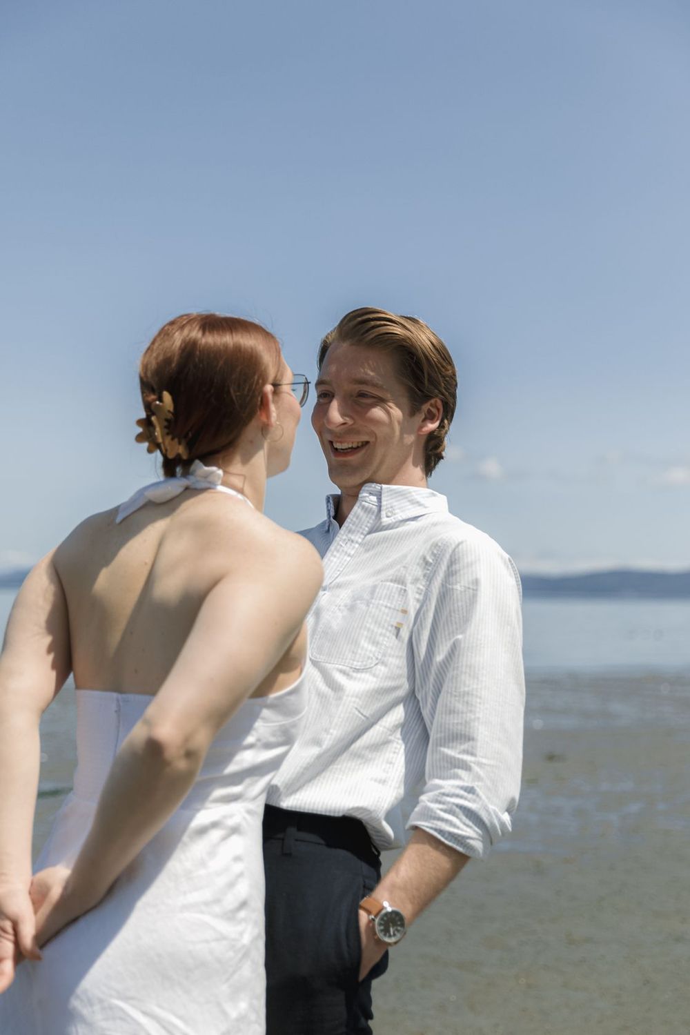 Smiling groom in white shirt holds hands with bride in white dress on a sunny beach.