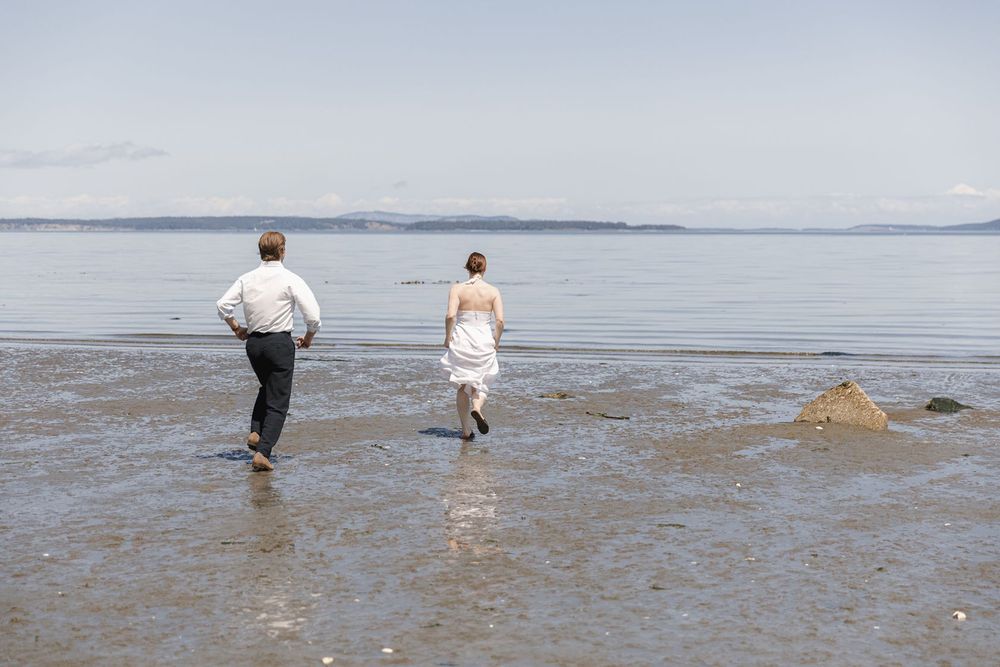 Bride and groom running joyfully along a scenic beach shoreline on their wedding day.