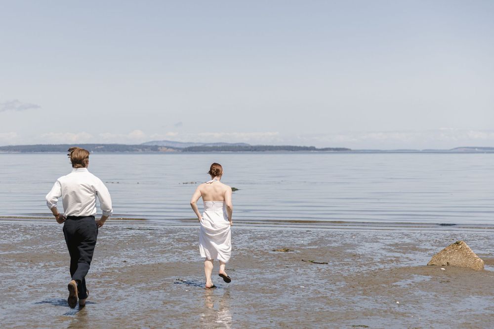 Couple in wedding attire running barefoot along a serene beach with mountain views.