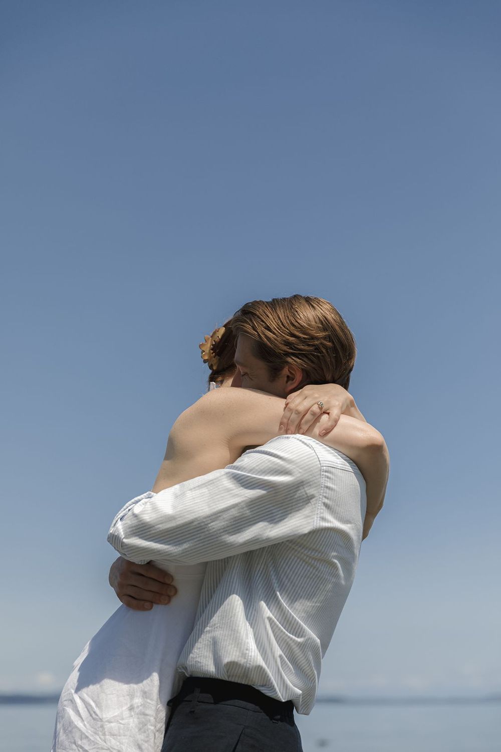 Couple embracing tenderly by the sea under a clear blue sky, man in white shirt holding woman close.