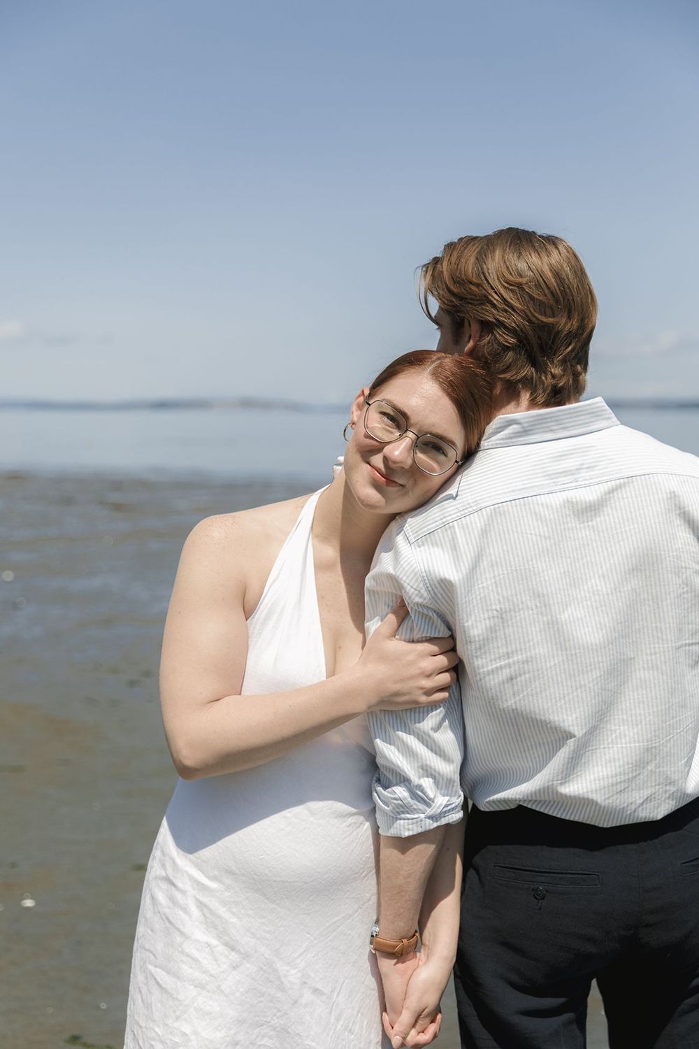 Bride in white halter dress embraces groom on beach, smiling over his shoulder with ocean backdrop.