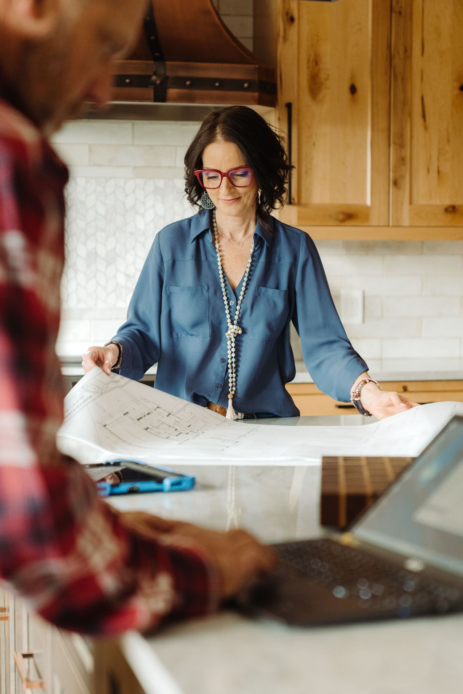Woman in blue blouse reviews architectural blueprints on kitchen counter with colleague holding laptop nearby.