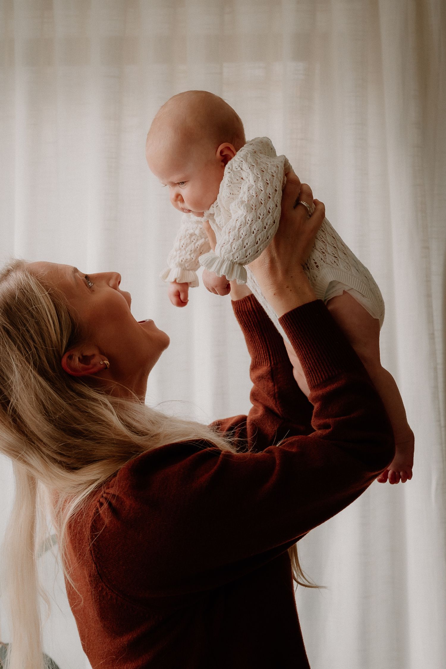 A blonde woman in a rust sweater lovingly lifts her smiling baby in a white knit outfit near a bright window.