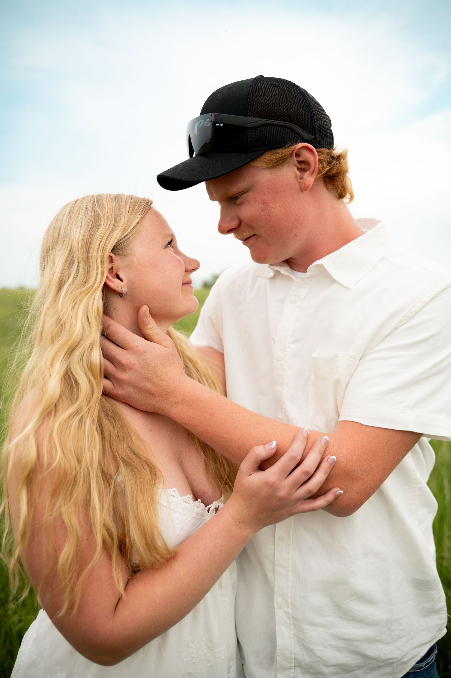 Young couple in white embracing in a green field, sharing a romantic moment under a cloudy sky.