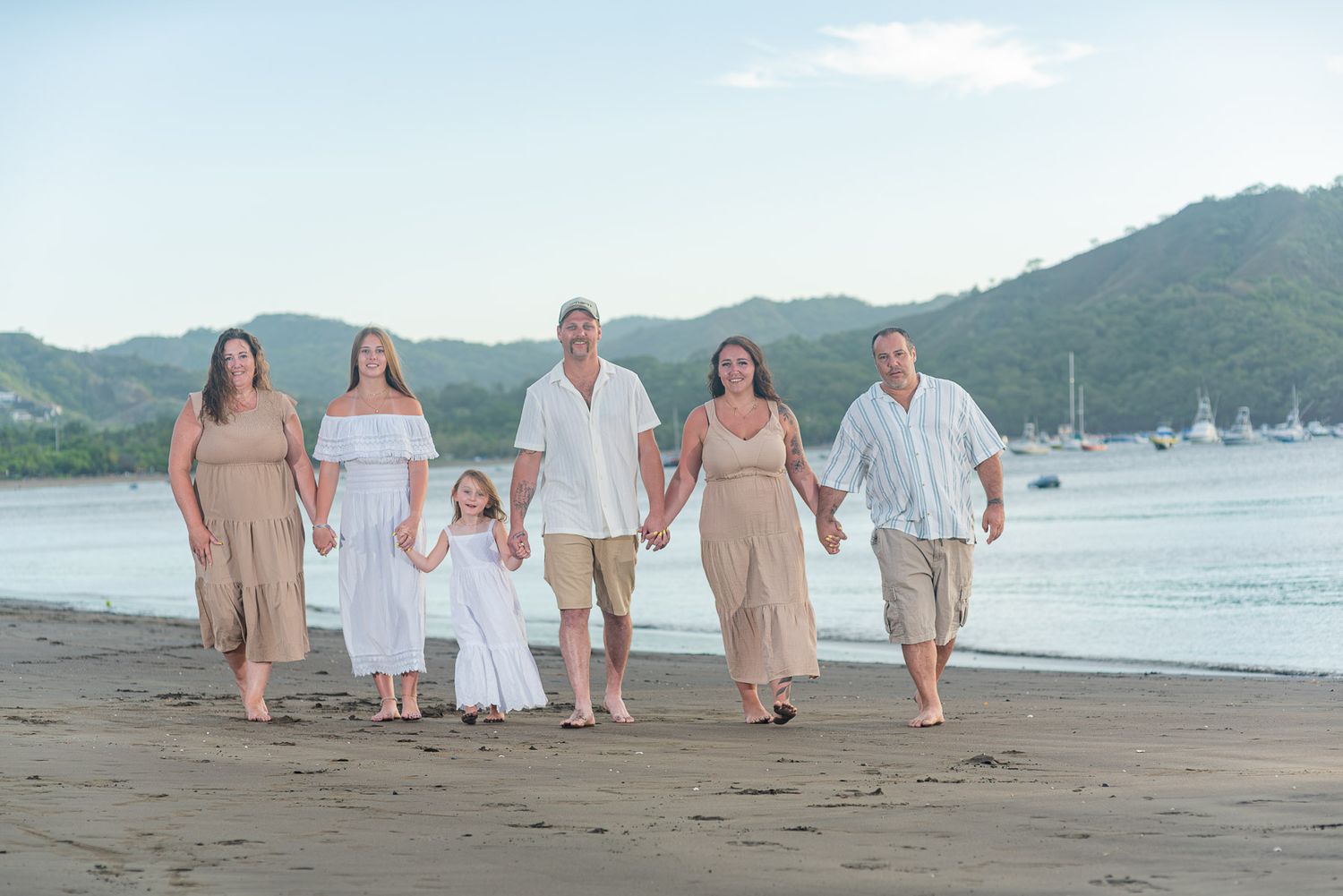 Family of six walking hand-in-hand on a tropical beach at sunset with lush green hills in the background.
