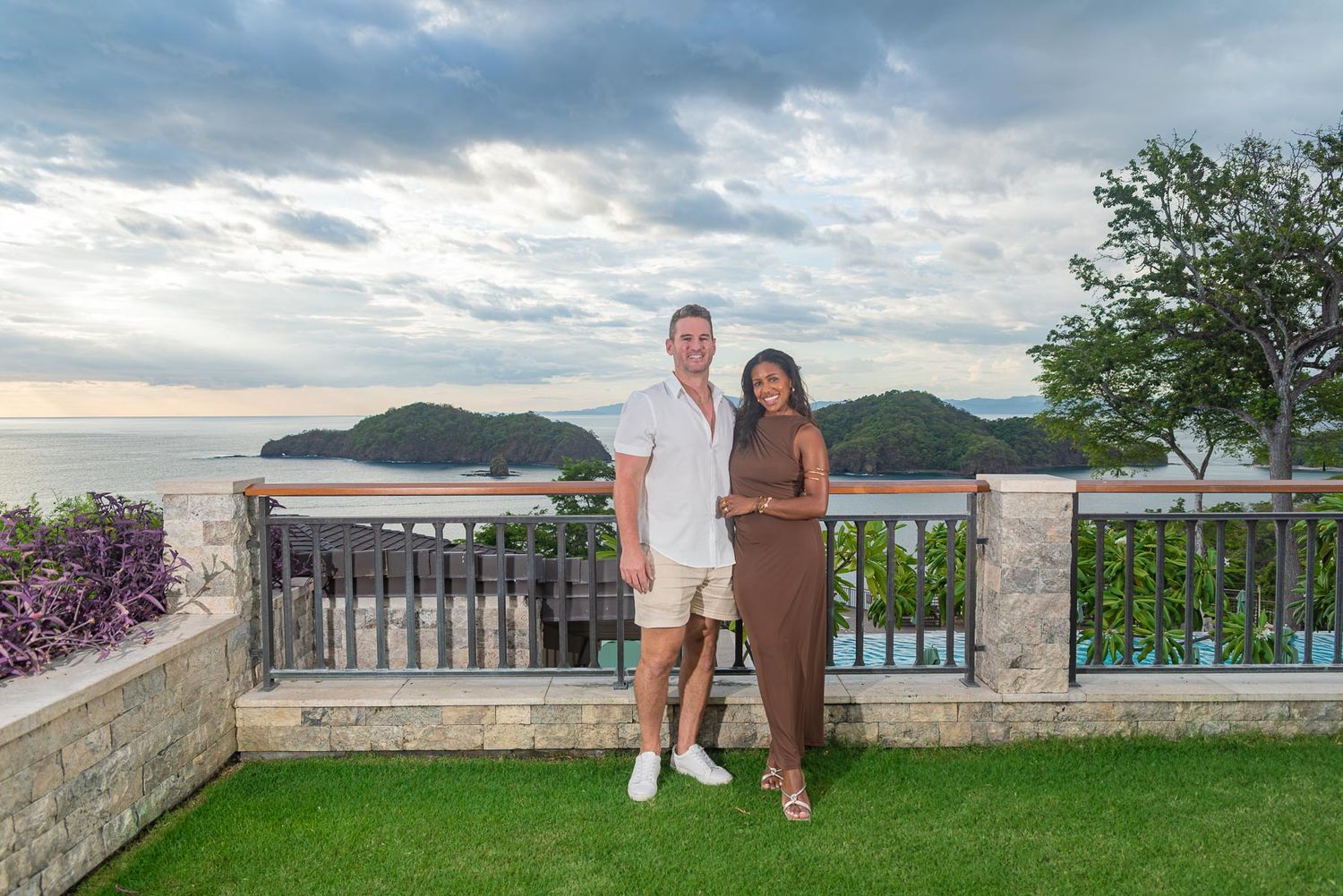 Couple posing on green lawn in front of white tropical resort with palm trees and cloudy sky.