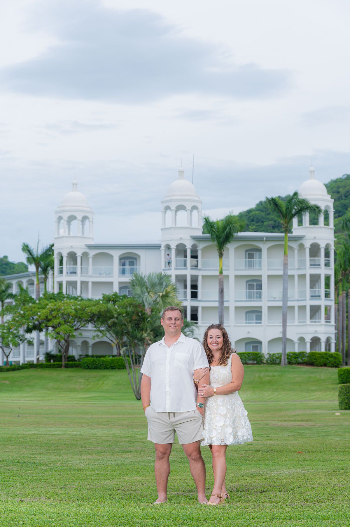 Couple posing on green lawn in front of white tropical resort with palm trees and cloudy sky.