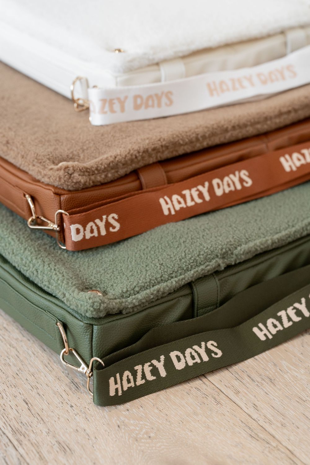 Stack of three Hazey Days pet beds in white, tan, and green with branded straps on wood floor.