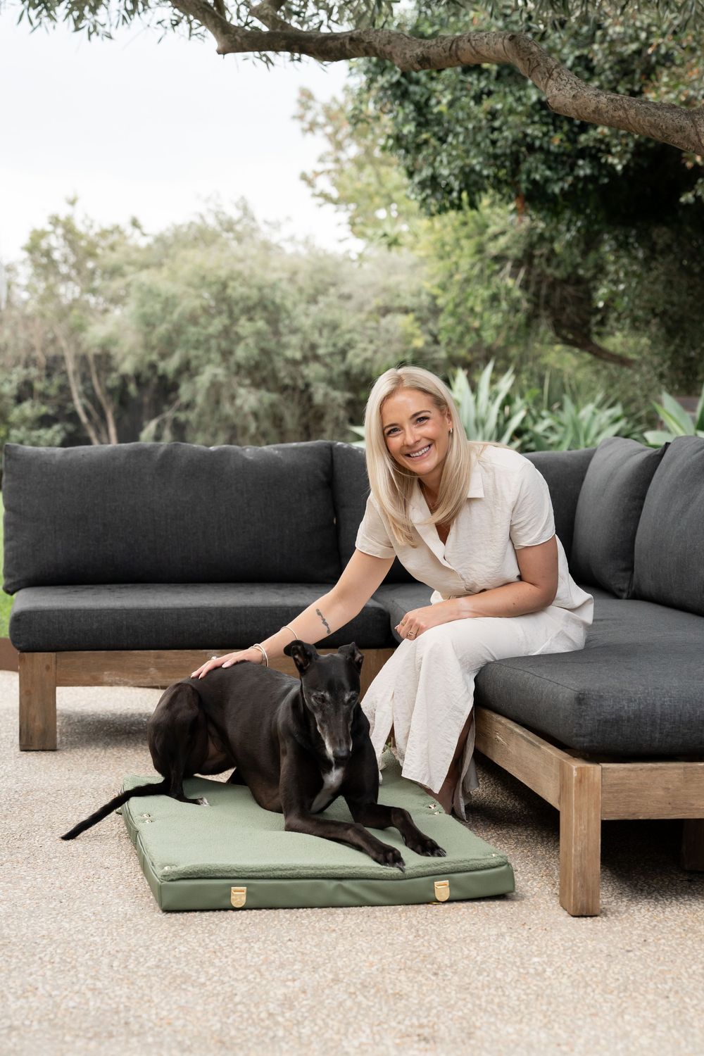 Smiling blonde woman sitting on dark outdoor sofa petting a black greyhound dog resting on a green Hazey Days pet bed.