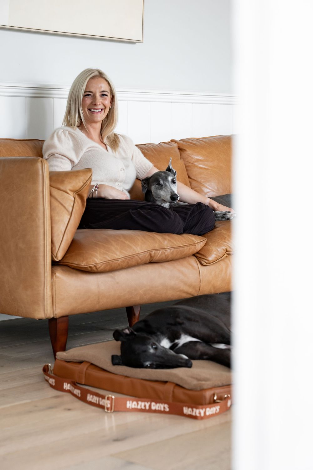 Smiling blonde woman relaxing on tan leather sofa with greyhound dog, another dog resting on Hazy Days pet bed below.