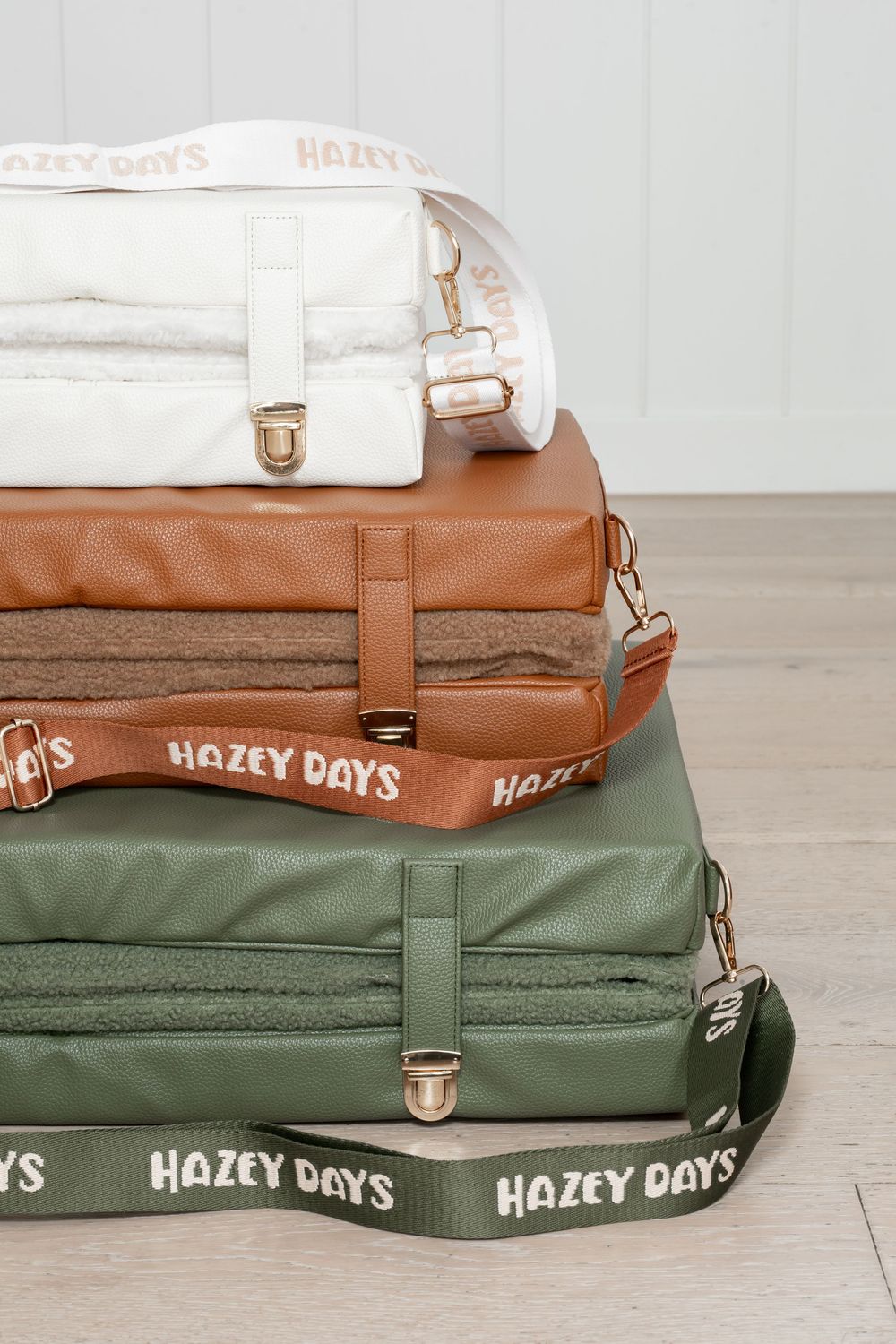 Stack of three Hazey Days dog beds in white, tan, and green colors with branded straps on wooden floor.
