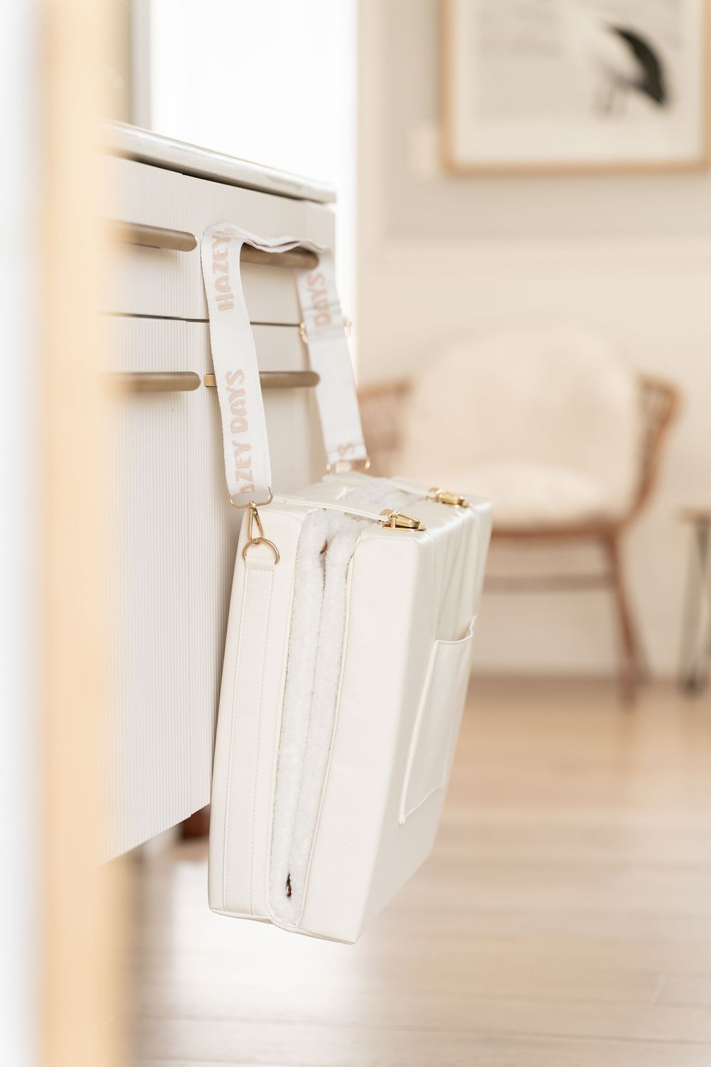 White leather handbag with gold hardware and branded strap hanging on a white cabinet in a bright, minimalist room.