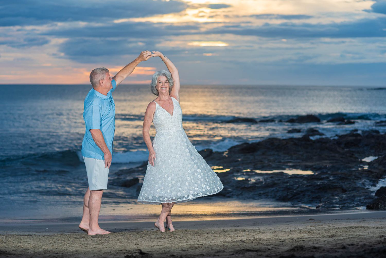 Elderly couple dancing romantically on a beach at sunset, woman in white dress being twirled by man in blue shirt.